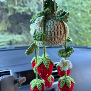 May include: A crocheted strawberry car charm. The charm has a beige crocheted pot with green leaves and white flowers. Red crocheted strawberries with green tops hang below, creating a decorative accessory for a vehicle.