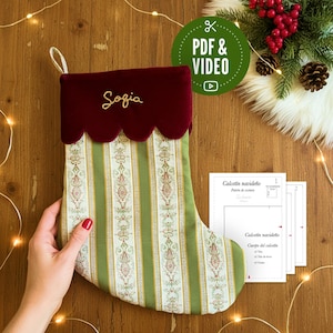 Puede incluir: Calcetín navideño con un estampado de rayas verdes y blancas, una parte superior festoneada burdeos y el nombre "Sofia" bordado en dorado. El calcetín está sostenido por una mano, con un icono de PDF y vídeo en el fondo.