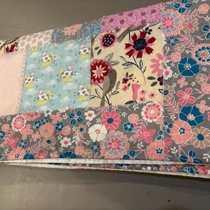 Könnte beinhalten: Ein Patchwork-Quilt mit verschiedenen Stoffmustern. Der Quilt hat einen hellrosa Abschnitt, einen blauen Abschnitt mit Kuhmotiven und florale Muster in Rosa, Blau und Grau. Der Quilt ist gefaltet und zeigt die verschiedenen Muster.