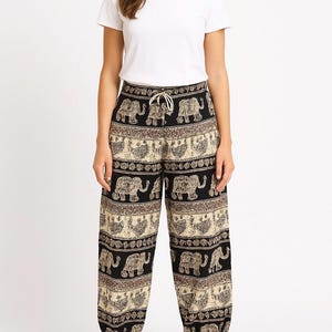 May include: Black and tan elephant print pants with a drawstring waist. The pants have a wide leg design and feature a repeating pattern of elephants and decorative borders. The pants are paired with a white t-shirt.