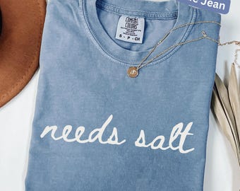 May include: A blue jean Comfort Colors t-shirt with the words "needs salt" in white script. A gold necklace with a pendant is draped across the shirt. The shirt is a small size.