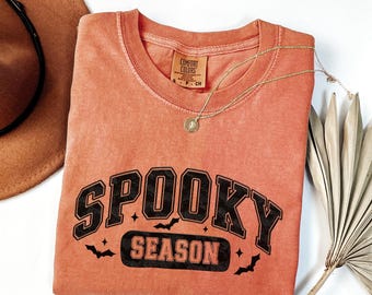 May include: An orange Comfort Colors t-shirt with the words "Spooky Season" in black block letters. The design includes bats and stars. A gold necklace with a pendant is draped over the shirt. A brown hat and dried palm leaf are in the background.