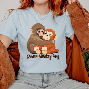 May include: Light blue t-shirt featuring a cartoon illustration of two hugging monkeys. One monkey is brown, the other is orange. The text "Punch Monkey Hug" is printed in brown script below the monkeys.
