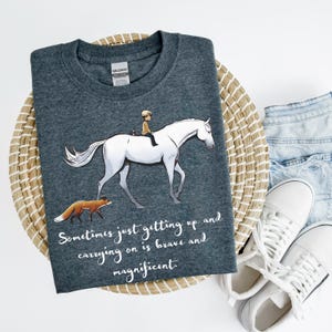 May include: Dark gray t-shirt featuring an illustration of a white horse with a rider and a fox. The shirt has the text "Sometimes just getting up and carrying on is brave and magnificent."