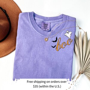 May include: A lavender Comfort Colors t-shirt with a Halloween design. The design includes the word "boo" in orange script, two black bats, and a white ghost. A gold necklace is draped across the shirt.