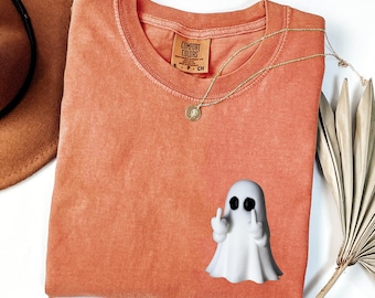 May include: Orange Comfort Colors t-shirt with a white ghost figurine giving the middle finger. A gold necklace with a pendant is on the shirt. The shirt is folded and next to a brown hat and a dried palm leaf.
