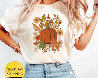 May include: A cream-coloured t-shirt with a detailed illustration of a pumpkin surrounded by autumn leaves and flowers in orange, brown, and green. The design has a fall or harvest theme. The words "NEXT DAY SHIPPING" are visible.