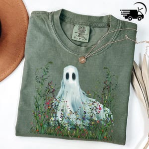 May include: An olive green Comfort Colors t-shirt featuring a ghost design surrounded by colorful wildflowers. The shirt has a crew neck and short sleeves. A delicate gold necklace with a pendant rests on the shirt. The shirt is labeled S-P-CH.