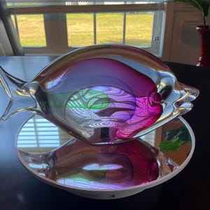 May include: A clear glass fish sculpture with internal swirls of pink, purple, and green, resting on a mirrored, white-rimmed dish. The fish has fins and a tail, reflecting light and colors.