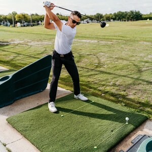 May include: A person swinging a golf club on a green artificial turf mat. The person is wearing a white shirt, black pants, sunglasses, and white shoes. A golf ball is on a tee, and other golf balls are on the mat.
