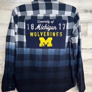 Könnte beinhalten: Ein blaues und weißes kariertes Flanellhemd mit einem Patch der University of Michigan auf der Rückseite. Der Patch zeigt das Logo der Schule, die Worte "University of Michigan", "WOLVERINES" und die Zahlen "18" und "17".