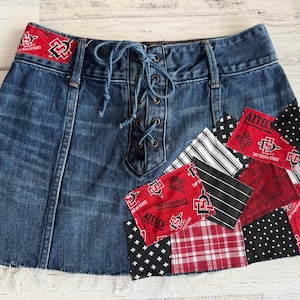 May include: Blue denim skirt with a lace-up front and patchwork accents. The patches feature red fabric with the text "AZTECS" and "San Diego State" along with various patterns, including polka dots and stripes.