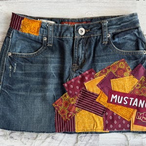 Op de afbeelding: Blauwe spijkerbroekrok met patchwork details. De rok heeft een rafelige zoom en een patch met het woord "MUSTANGS" in het wit. Het patchwork bevat bordeauxrode, gouden en bedrukte stoffen.