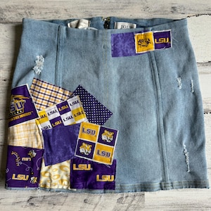 May include: Light blue denim skirt with patches featuring the LSU logo and colors. Patches include purple, gold, and white squares, polka dots, and plaid patterns. The skirt has a zipper closure and distressed details.