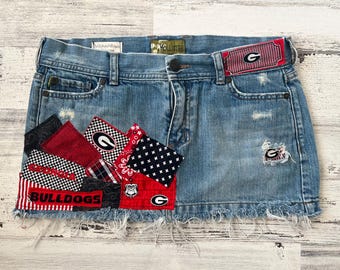 May include: Light blue denim skirt with distressed details and a frayed hem. Patches of red, black, and white fabric with various patterns and the word "BULLDOGS" are sewn onto the skirt. A red and white striped belt loop features a "G" logo.