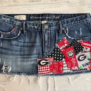 Puede incluir: Minifalda vaquera desgastada con parches con el logotipo y los colores de los Bulldogs de la Universidad de Georgia. La falda tiene un dobladillo deshilachado, detalles rasgados y el logotipo de Abercrombie and Fitch.