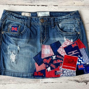 Pode incluir: Uma saia de ganga com remendos de vários tecidos, incluindo vermelho, branco e azul. Os remendos apresentam as palavras "Ole Miss" e "Rebel" e o logotipo da Universidade do Mississippi.