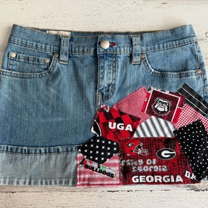 Puede incluir: Minifalda vaquera con parches de tela roja, negra y blanca. Los parches presentan los textos "UGA", "Bulldogs" y "University of Georgia", junto con el logotipo de los Georgia Bulldogs.