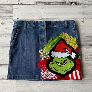 Peut inclure: Une jupe en jean bleu avec un motif patchwork mettant en scène un personnage vert du Grinch portant un bonnet de Père Noël rouge. Le patchwork comprend des tissus rouges, verts, blancs et noirs.