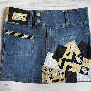 Puede incluir: Falda vaquera azul con parches UCF. La falda presenta un parche UCF negro y dorado en la cintura y el bolsillo, y múltiples parches con el logotipo UCF y el texto Knights. La falda tiene dos botones y un bolsillo.