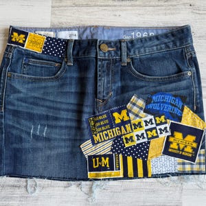 May include: A blue denim skirt with patches featuring the University of Michigan logo and text. The skirt has a raw hem and distressed details. The waistband includes the text "Gap 1969" and the number "27/4".