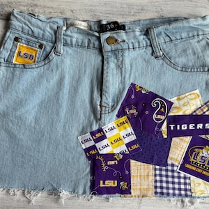 Pode incluir: Saia jeans azul claro com bainha crua. Apresenta um logotipo LSU amarelo e roxo no bolso frontal e patchwork com a palavra "Tigers" e logotipos LSU. O tamanho da saia é de 91 cm.