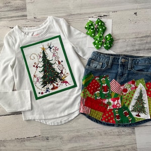 May include: A white long-sleeved shirt featuring a green framed image of a Christmas tree with cartoon characters decorating it. A denim skirt with a patchwork design of red, green and white fabric.