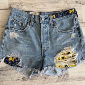 May include: Light blue denim shorts with distressed details. The waistband features a yellow and blue pattern with the letter "M". Patches of the same pattern are visible under the distressed areas of the shorts.