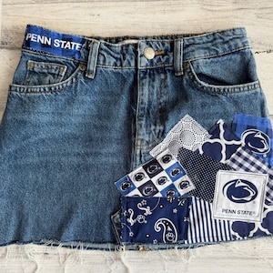 May include: Blue denim skirt with a raw hem and a waistband that reads "PENN STATE". Patches of patterned fabric in blue and white are sewn onto the skirt, featuring the Penn State logo and various designs.