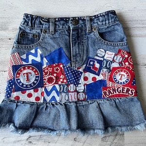 May include: A denim skirt with a ruffled hem, featuring a patchwork design of blue, red, and white fabric. The patches include baseball-themed designs, including the Texas Rangers logo.