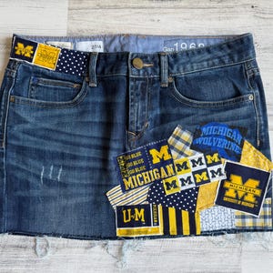 Op de afbeelding: Blauwe spijkerbroekrok met distressed details en patches met het logo en de tekst van de Universiteit van Michigan. De rok heeft een rafelige zoom en een knoopsluiting. De tailleband heeft de tekst "Gap 1969" en de maat "27/4".