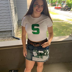 Op de afbeelding: Een vrouw draagt een wit T-shirt met een groene glitter "S" op de voorkant en een spijkerrok met Michigan State patches.