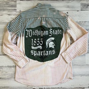 May include: A white and pink plaid button-up shirt with a green patch on the back that says "Michigan State 1855 Founded Spartans".