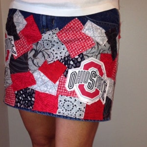 Pode incluir: Uma saia de ganga com um design de patchwork com quadrados de tecido vermelho, branco e preto. A saia tem um grande remendo com o logotipo do Ohio State na frente.