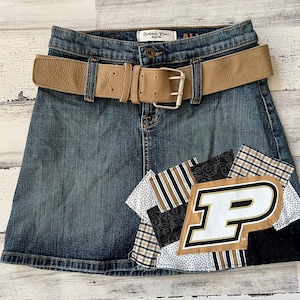 May include: A blue denim skirt with a brown leather belt and a patchwork design featuring a white letter "P" on a black and gold background.