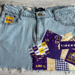 May include: Light blue denim skirt with a raw hem. Features a yellow and purple LSU logo on the front pocket and patchwork with the word "Tigers" and LSU logos. The skirt size is 36 inches.
