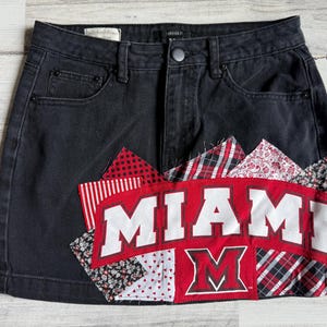 Pode incluir: Saia jeans preta com design de patch. O patch apresenta a palavra "MIAMI" em letras brancas sobre um fundo vermelho, com um logotipo "M" vermelho. O patch é cercado por vários padrões de tecido em vermelho, branco e preto.