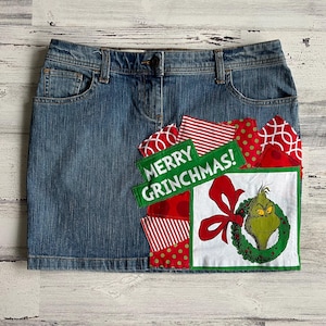 Op de afbeelding: Een denim rok met een patchwork ontwerp met rode, groene en witte stof. De stof bevat de woorden "Merry Grinchmas!" en een cartoon afbeelding van de Grinch.