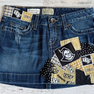 May include: A denim mini skirt with patches featuring the UCF Knights logo in black, gold, and white. The skirt has a button closure, pockets, and distressed details. The patches are sewn onto the skirt.