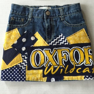 Könnte beinhalten: Ein Jeansrock mit Patches aus gelbem und blauem Stoff. Die Patches haben Punkte, Zickzackmuster und die Worte "Oxford Wildcats".
