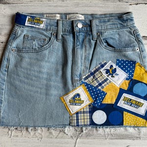 Pode incluir: Minissaia jeans azul claro com bainha crua e detalhes de patchwork. O patchwork inclui tecidos azuis, amarelos e brancos com o texto "DELAWARE BLUE HENS" e um logotipo de uma galinha azul.
