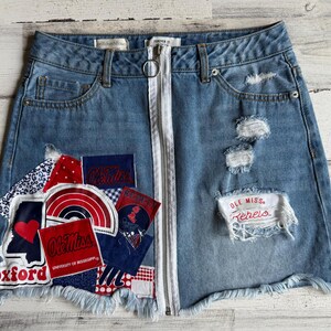 Könnte beinhalten: Blaue Jeans-Minirock mit Distressed-Details und weißem Reißverschluss. Auf der Vorderseite sind Aufnäher mit roten, weißen und blauen Designs sowie den Texten "Oxford" und "Ole Miss Rebels" aufgenäht. Der Rock hat einen ausgefransten Saum.