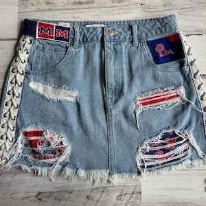 Può includere: Gonna di jeans azzurra con dettagli strappati, con toppe con testo "MM" e "Ole Miss". La gonna ha lati con lacci e bordi sfilacciati, con accenti rossi e bianchi.