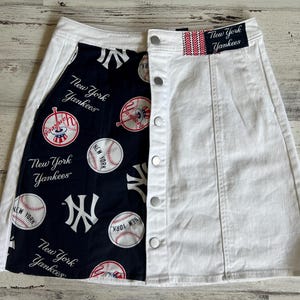 Könnte beinhalten: Ein weißer Jeansrock mit Knopfleiste vorne und einem schwarzen und weißen Stoffpanel mit New York Yankees-Motiv auf der linken Seite. Das Stoffpanel zeigt Baseballs, Schläger und das New York Yankees-Logo.