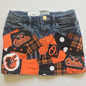 May include: A denim skirt with patches featuring the Baltimore Orioles logo, a bird, and the text "Orioles" in orange and black. The patches are arranged in a patchwork design with black and white polka dots, plaid, and orange fabric.
