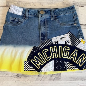Puede incluir: Una falda vaquera con un efecto degradado amarillo y un diseño de patchwork con la palabra "MICHIGAN" en letras amarillas. El patchwork incluye una tela blanca con la palabra "MICHIGAN" impresa en negro.