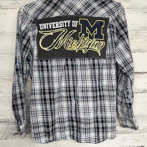 Könnte beinhalten: Ein blaues und weißes kariertes Flanellhemd mit einem grauen Patch auf der Rückseite, auf dem "University of Michigan" mit einem gelben "M" und einer gelben Schrift steht.