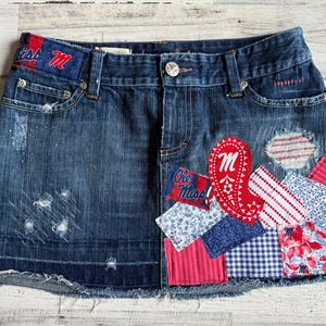 Può includere: Gonna di jeans blu con dettagli strappati e toppe decorative. Le toppe includono motivi paisley, floreali e a righe, insieme al testo "Ole Miss" e alla lettera "M". La gonna ha un orlo sfilacciato.