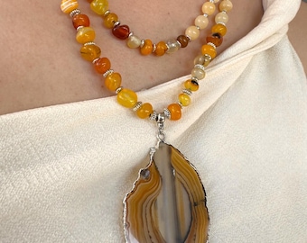 Puede incluir: Un collar de varias capas con un colgante grande de ágata pulida con vetas marrones y blancas, acentuado por un borde de metal plateado. El collar incluye hilos de cuentas de piedras preciosas amarillas y naranjas, creando una paleta de colores cálidos y terrosos.