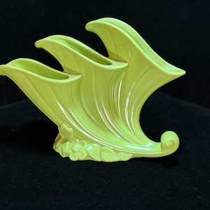 May include: A lime green ceramic vase featuring three distinct, upward-curving, horn-shaped openings. The base of the vase is decorated with a molded relief pattern of small flowers and leaves, and the structure ends in a curled scroll detail.